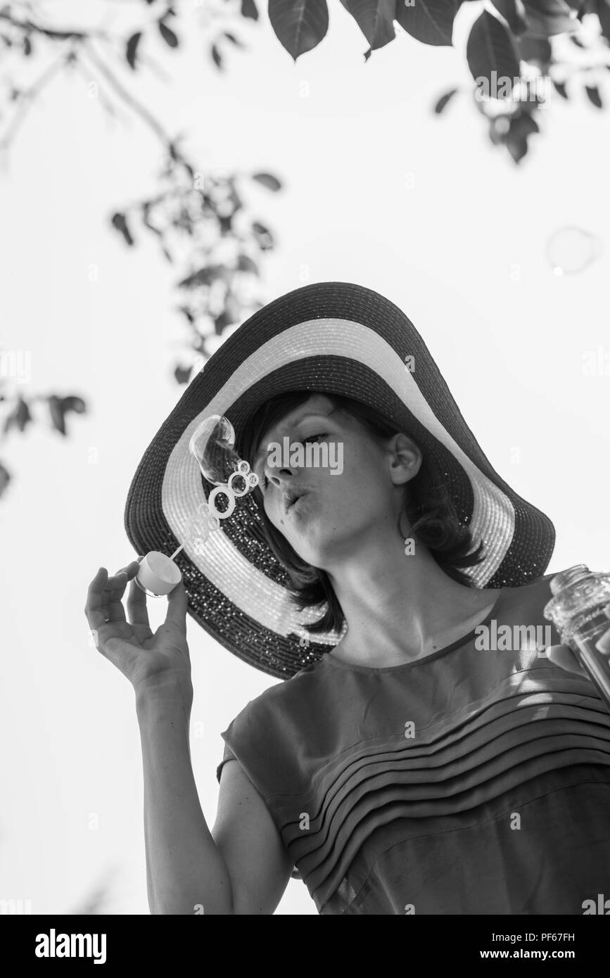 Scala di grigi ritratto di una donna attraente in una vasta colmato cappello a soffiare bolle all'aperto nel giardino all'ombra di un albero. Foto Stock