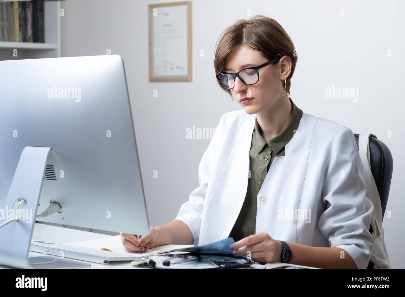 Praticante femmina lavora al moderno medico ufficio. Giovane medico prendendo appunti al lavoro nella parte anteriore del computer desktop Foto Stock