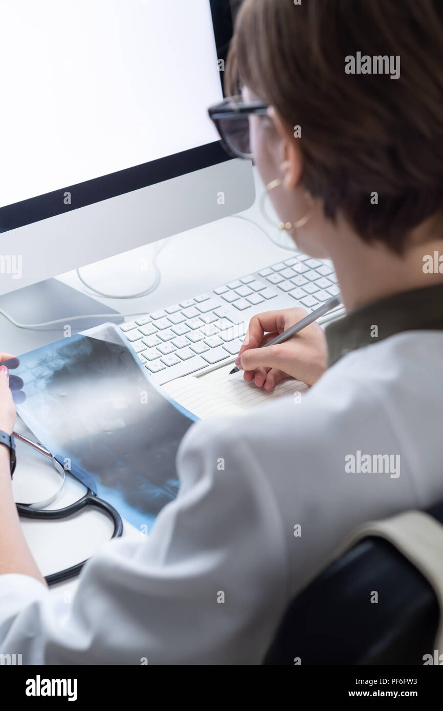 Praticante femmina lavora al moderno medico ufficio. Giovane medico prendendo appunti al lavoro nella parte anteriore del computer desktop Foto Stock
