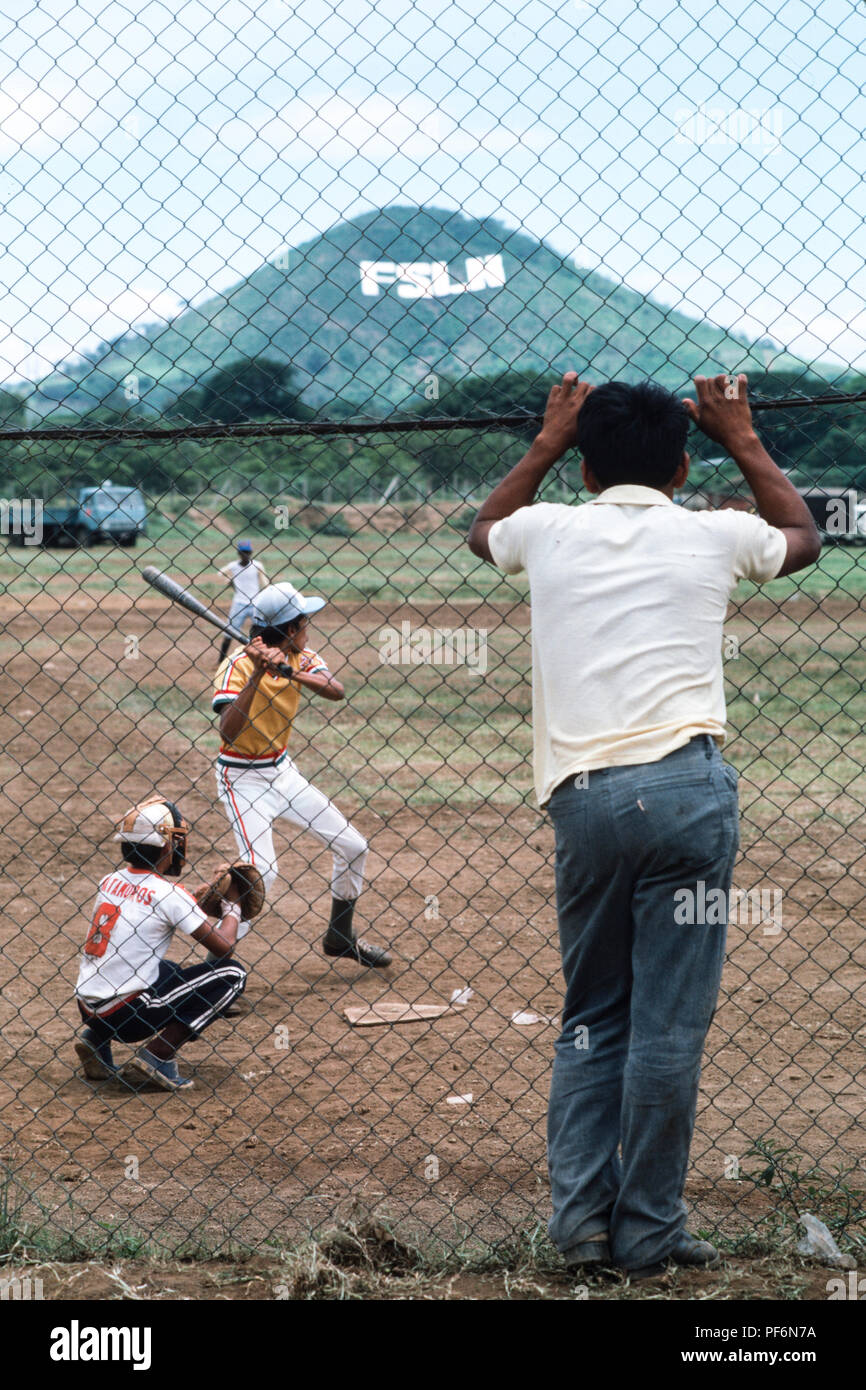 Managua, Nicaragua, giugno 1986; la gente a giocare a baseball. La segnaletica del Sandinista FSLN è visibile a grandi lettere sulla collina opposta. Foto Stock