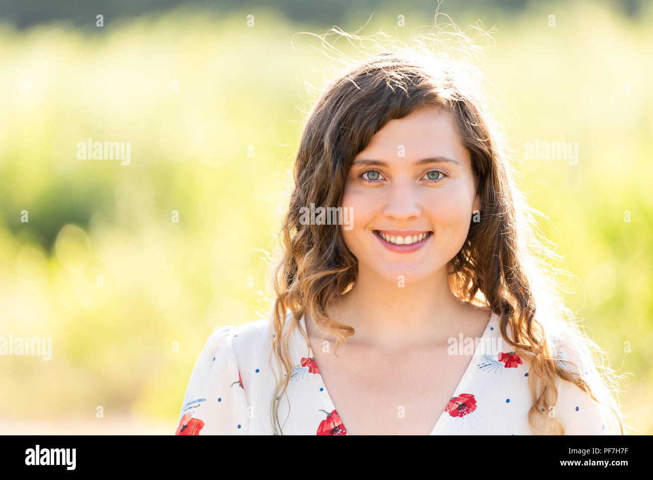 Closeup Ritratto di giovane volto di donna sorridente felice nella soleggiata estate parco verde al tramonto che mostra denti, capelli mossi nel vento, sfondo bokeh di fondo Foto Stock