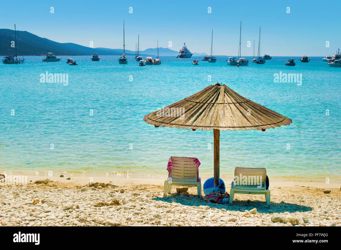 Uno giallo paglierino con ombrellone e due sedie a sdraio sulla sabbia ghiaiosa spiaggia. Molti yacht nel blu mare azzurro laguna in una baia vicino alla costa di Isola. Blu Foto Stock