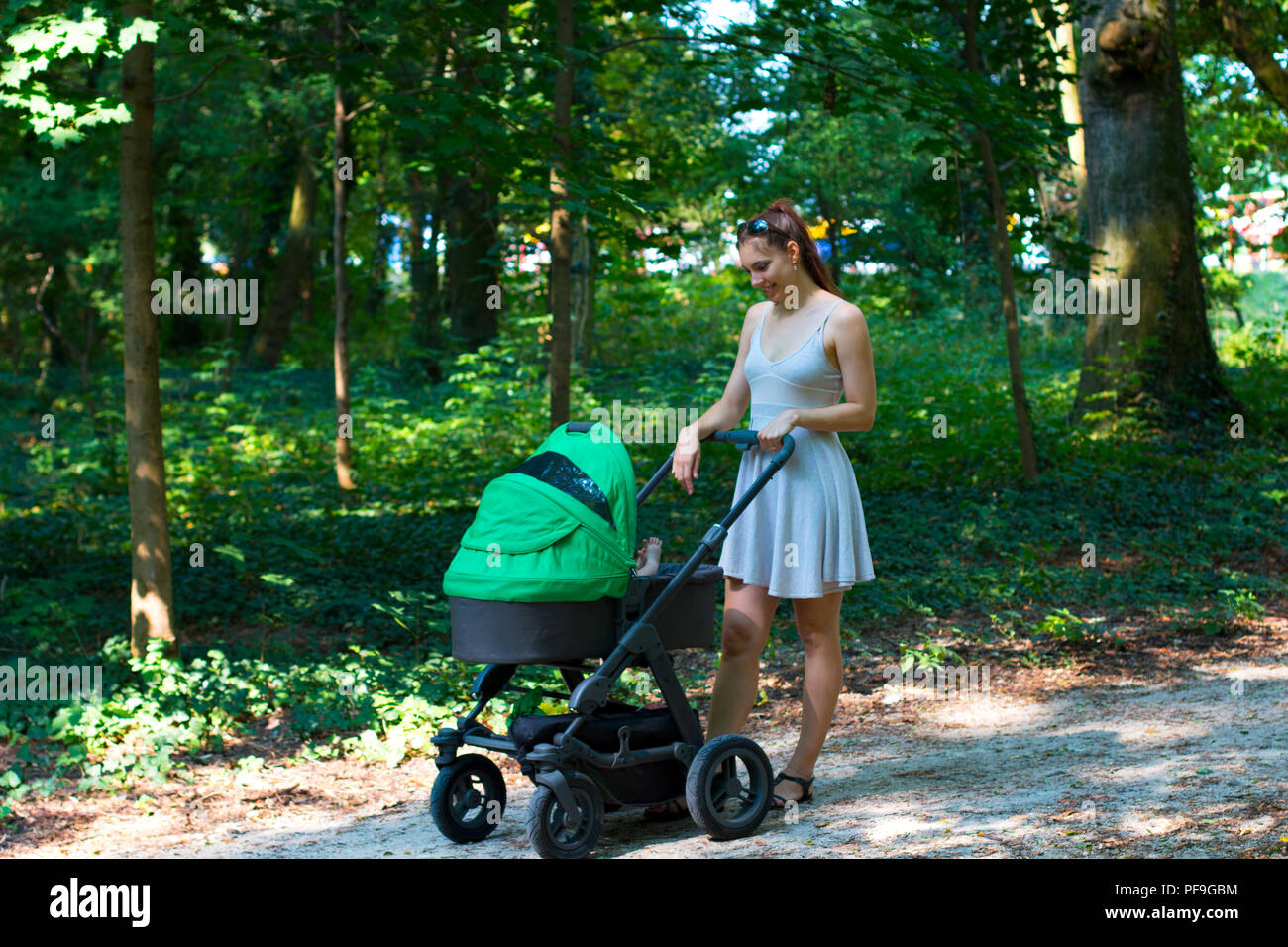 La natura a piedi con il passeggino, giovane madre nel bel vestito camminando sul marciapiede della foresta con il suo bambino nel passeggino, godendo di aria fresca e sorridente Foto Stock