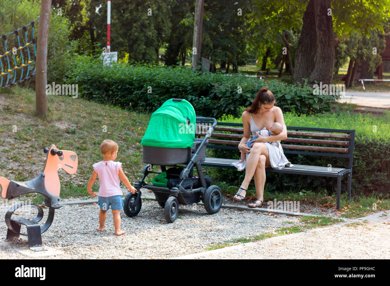 La maternità, giovane bella madre sul congedo parentale allattare al seno il suo bambino nel parco giochi per bambini, curioso bambino sta guardando a lei, la madre e il bambino Foto Stock