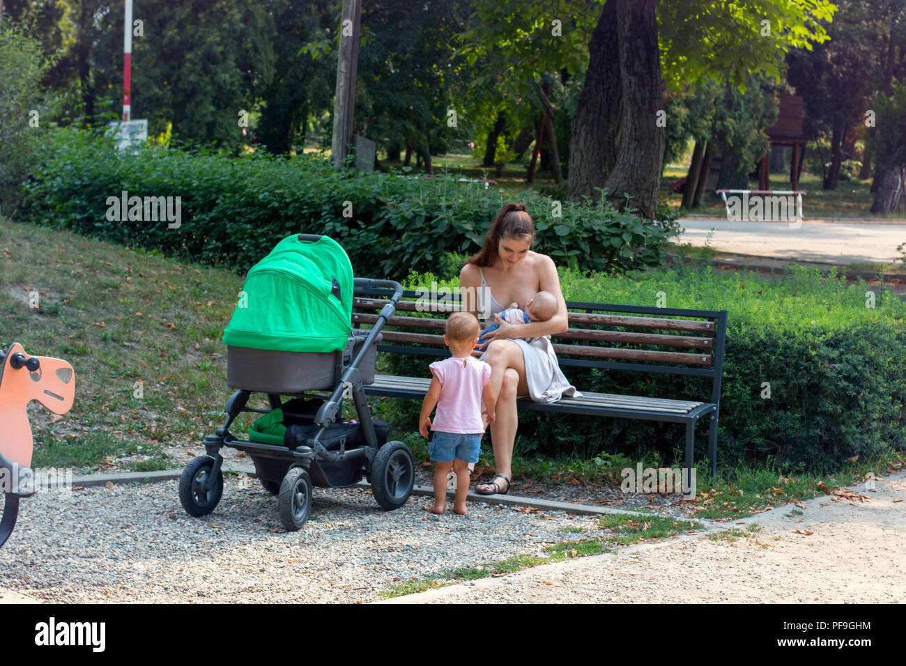 Happy mom sul congedo di maternità è fuori con i suoi bambini, giovani donne madre allattamento al seno nel parco, mentre curioso bambino sta cercando su di essi Foto Stock