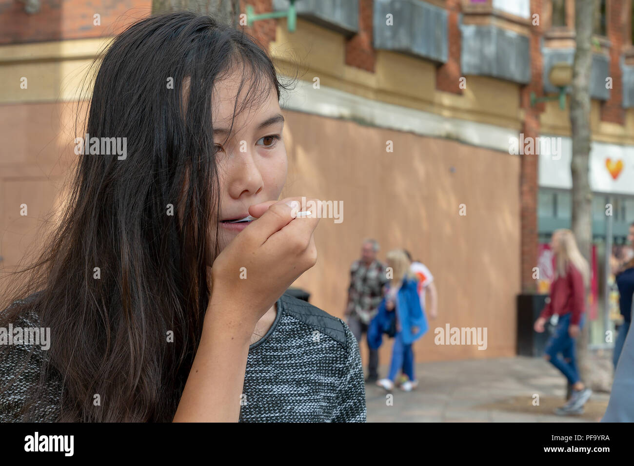 Inglese/Thai ragazza adolescente con lunghi capelli neri mangia una granita blu nel centro citta' di Warrington Foto Stock