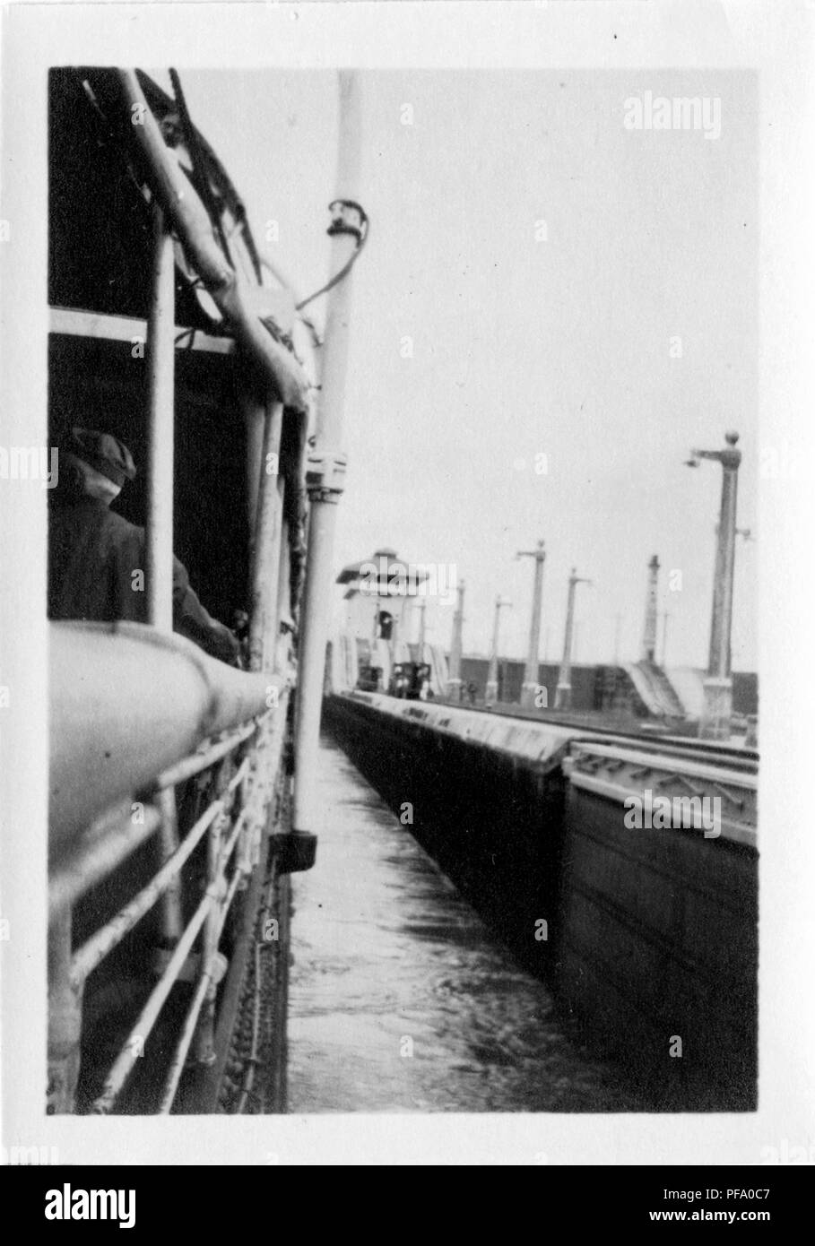 Fotografia in bianco e nero, che mostra la parte posteriore di un uomo in piedi vicino alla ringhiera di una nave che si trova all'interno di un canale di Panama, serratura con luce elettrica poli, Panama Canal di binari ferroviari, e di un blocco della torre di controllo visibile su la sponda del Canal Grande a destra, 1915. () Foto Stock