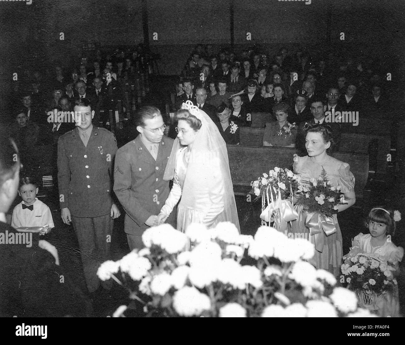 Fotografia in bianco e nero, inquadratura da dietro la spalla destra di un officiante del matrimonio, che mostra un groom in uniforme militare, in piedi accanto ad una sposa che indossa un abito bianco, velo, tiara, e i bicchieri con il miglior uomo (anche in uniforme) un giovane portatore di anello, la cameriera di onore, tenendo fiori, una giovane damigella holding di fiori e di un gran numero di ospiti seduti in banchi sullo sfondo, fotografato in Ohio, 1945. () Foto Stock