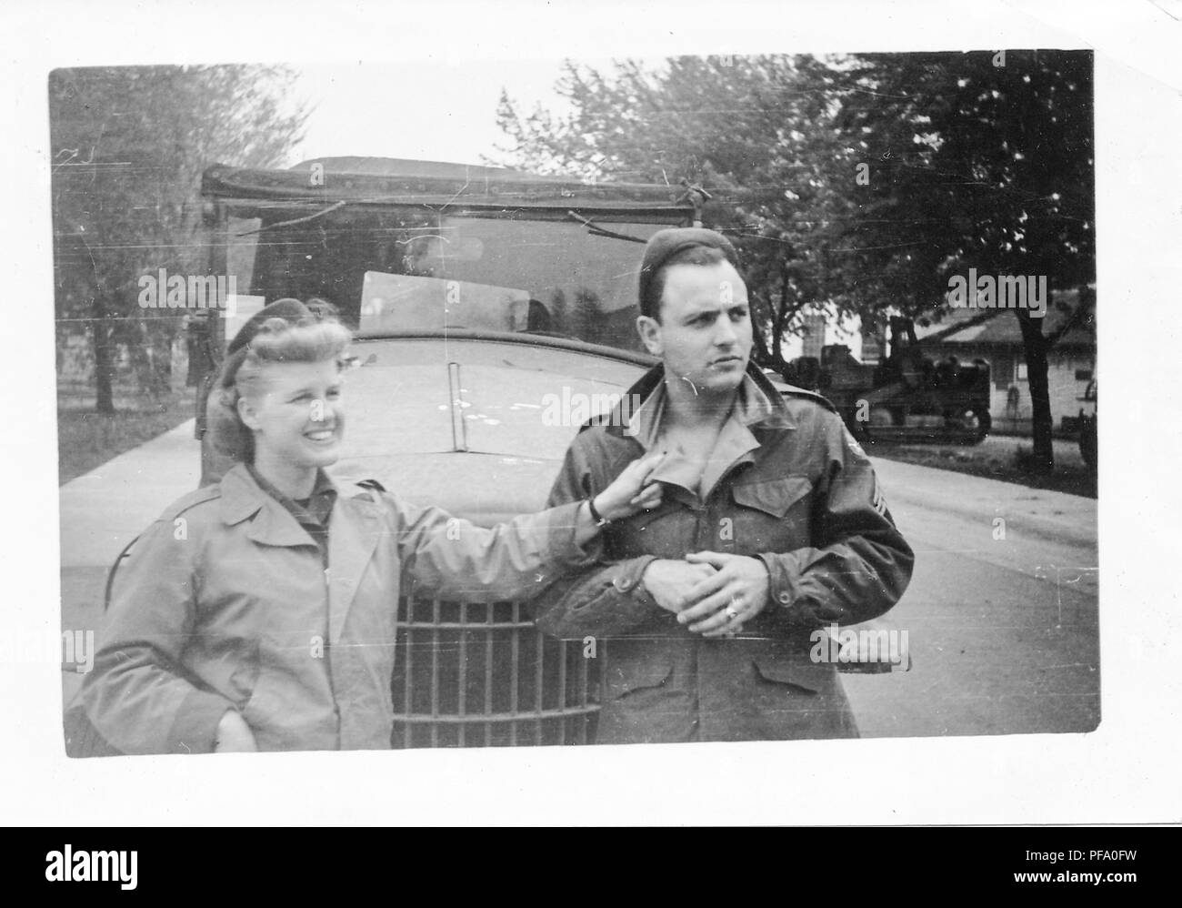 Fotografia in bianco e nero, mostrando un uomo e una donna, dalla vita in su, entrambi indossano uniformi militari e in piedi di fronte a un soft-top veicolo, la donna è sempre sorridente e la parte di afferramento dell'uomo collare con una mano tesa, entrambi guardano a sinistra della videocamera, con alberi, edifici e un altro veicolo in background, probabilmente fotografato durante la II Guerra Mondiale, 1945. () Foto Stock