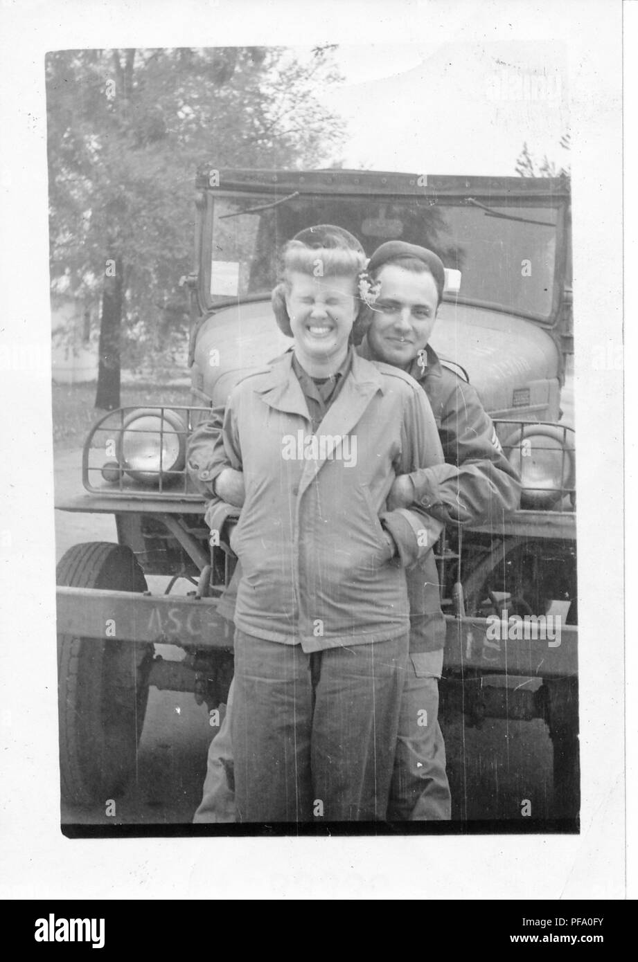 Fotografia in bianco e nero, mostrando un uomo dietro una donna, entrambi in tre quarti di lunghezza vista, indossano uniformi militari e in piedi di fronte a un soft-top veicolo, l'uomo ha le braccia legate attraverso la donna di braccia e sorrisi direttamente alla fotocamera, la donna ha il suo mani in tasca, i suoi occhi chiusi ermeticamente e rende un volto alla fotocamera, probabile fotografato durante la II Guerra Mondiale, 1945. () Foto Stock