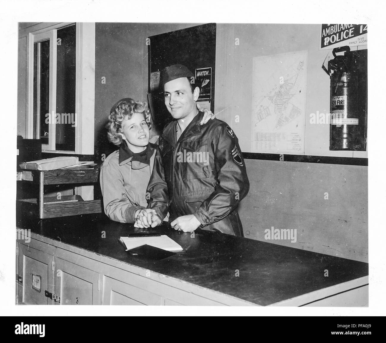 Fotografia in bianco e nero, che mostra un uomo in piedi accanto a una bionda donna, entrambi mostrati dalla vita in su, indossano uniformi militari e di stare al chiuso, dietro un lungo bancone, con un estintore in background, l'uomo sorrisi a qualcosa al di fuori della telecamera mentre la donna si affaccia la telecamera, loro legato le mani a destra il resto sul bancone mentre la donna di braccio sinistro avvolge l'uomo della spalla, probabile fotografato durante la II Guerra Mondiale, 1945. () Foto Stock
