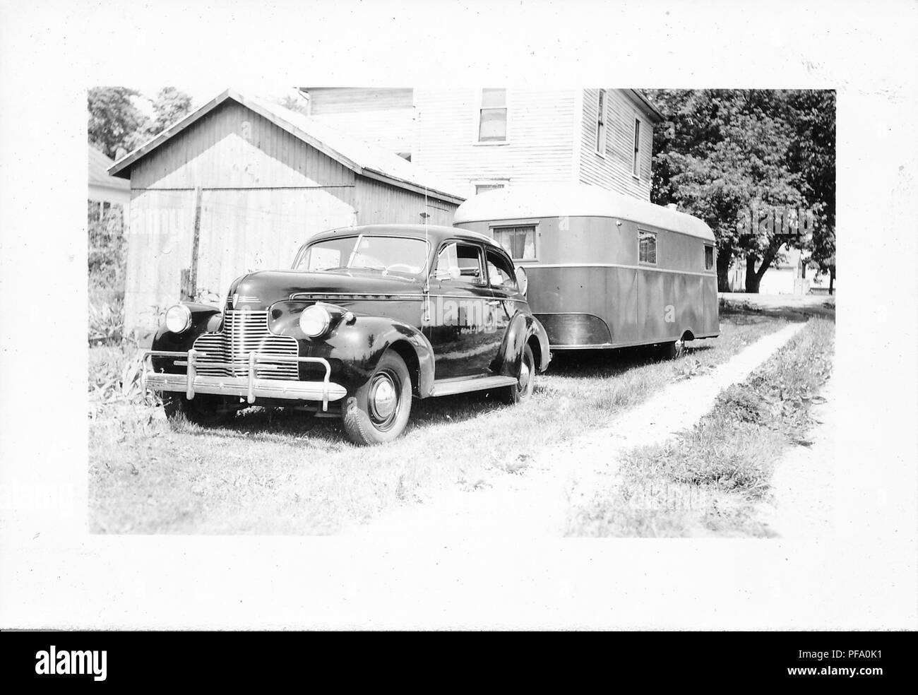 Fotografia in bianco e nero, che mostra la parte posteriore di un lucido, scuro, vintage, Chevrolet berlina, con una roulotte rimorchio attaccato al suo retro, parcheggiato all'esterno accanto ad una casa in legno e versato, probabilmente fotografato in Ohio nel decennio dopo la II Guerra Mondiale, 1950. () Foto Stock