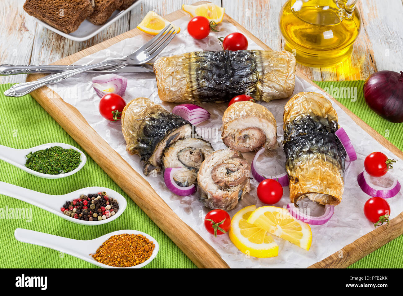 Un sano cotto i filetti di sgombro in rotoli, i pomodori e le fette di limone su carta pergamena bianca sul tagliere con coltello e forchetta, spezie, pane di segale Foto Stock