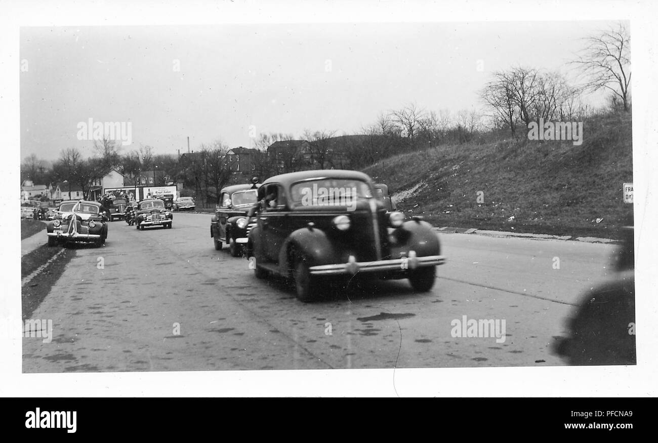 Fotografia in bianco e nero che mostra diverse auto d'epoca, compreso un buio, Chevrolet berlina a metà-centro, come guidare su una strada trafficata, verso la fotocamera, con case residenziali, un terrapieno e alberi sfrondato in background, probabilmente fotografato in Ohio nel decennio dopo la II Guerra Mondiale, 1945. () Foto Stock