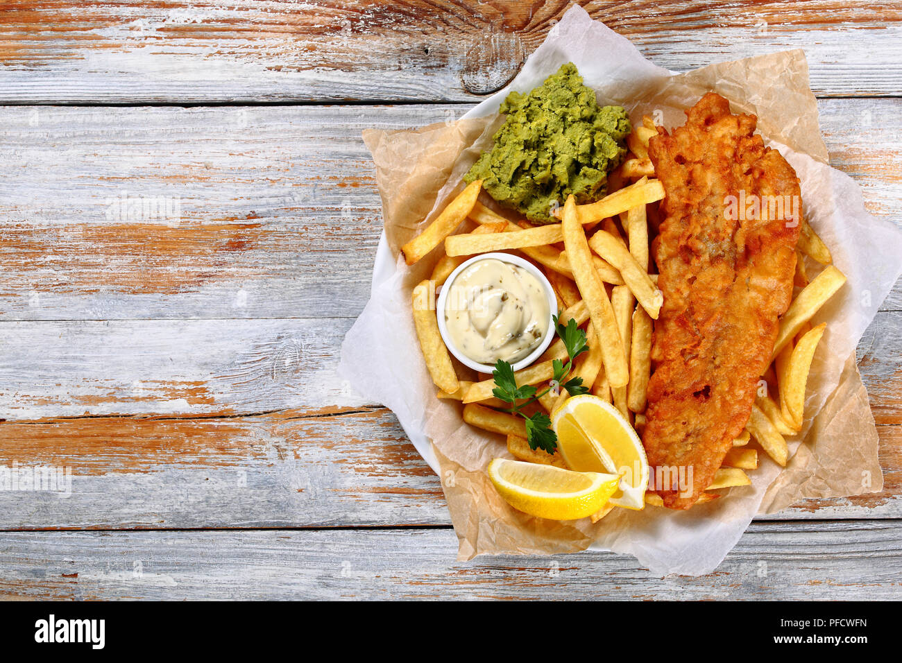 Delizioso croccante di pesce e patatine fritte - cod, patatine fritte, le fette di limone, salsa tartara e purea di piselli su piastra su carta sul vecchio tavolo in legno, authen Foto Stock