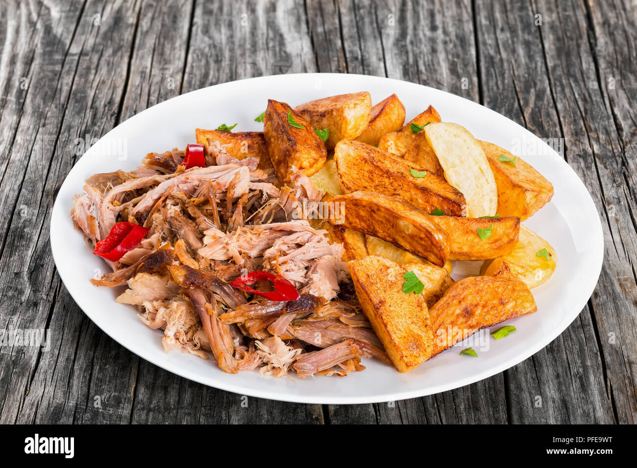 Porzione di tirata slow-cotta deliziosa carne arrostita al forno con patate fritte cunei cosparsa di prezzemolo sulla piastra bianca sul legno scuro, sfondo Foto Stock