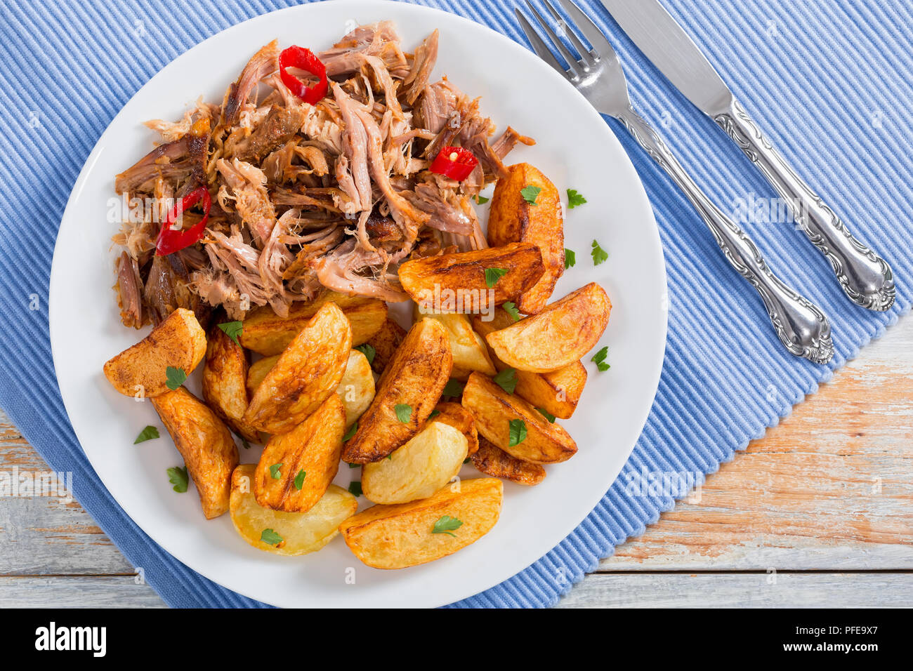 Tirato slow-cotto spalla di maiale grigliato in forno con patate fritte cunei, peperoncino e spezie sul lilla tabella MAT con coltello e forchetta, su legno t Foto Stock