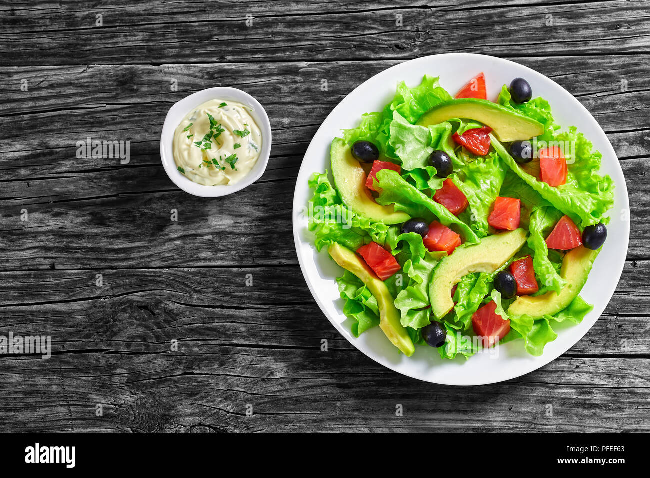 Deliziosa insalata fresca di foglie di lattuga, fette di avocado, pezzetti di salmone salato e olive nere su piastra bianca sul tavolo di legno con fatti in casa mayo Foto Stock