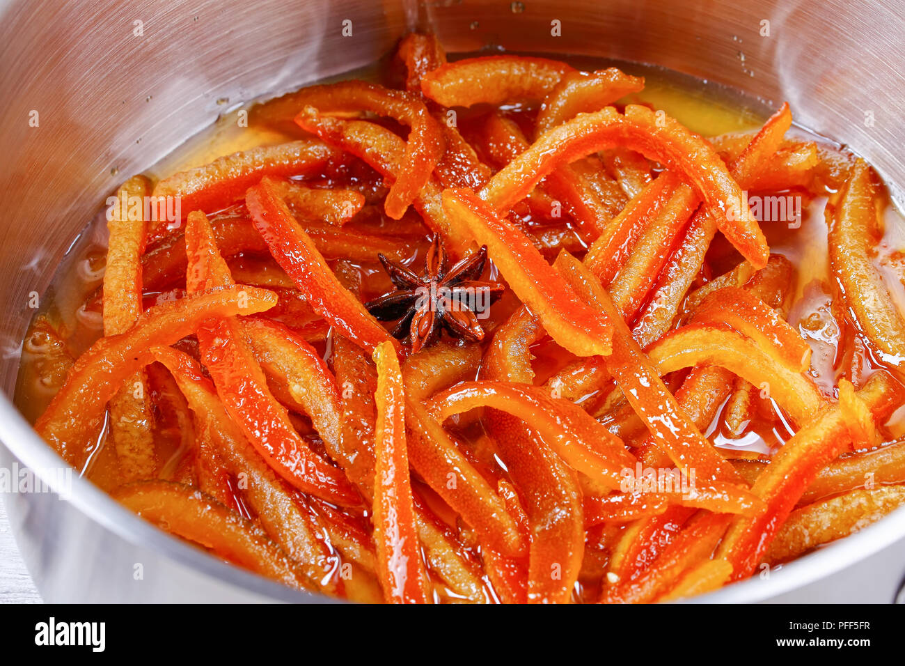 Deliziosi dolci fatti in casa arancia candita e bucce di limone cotte in sciroppo bollente con la cannella e la noce moscata e anice stella nella pentola, vista da sopra, macro Foto Stock