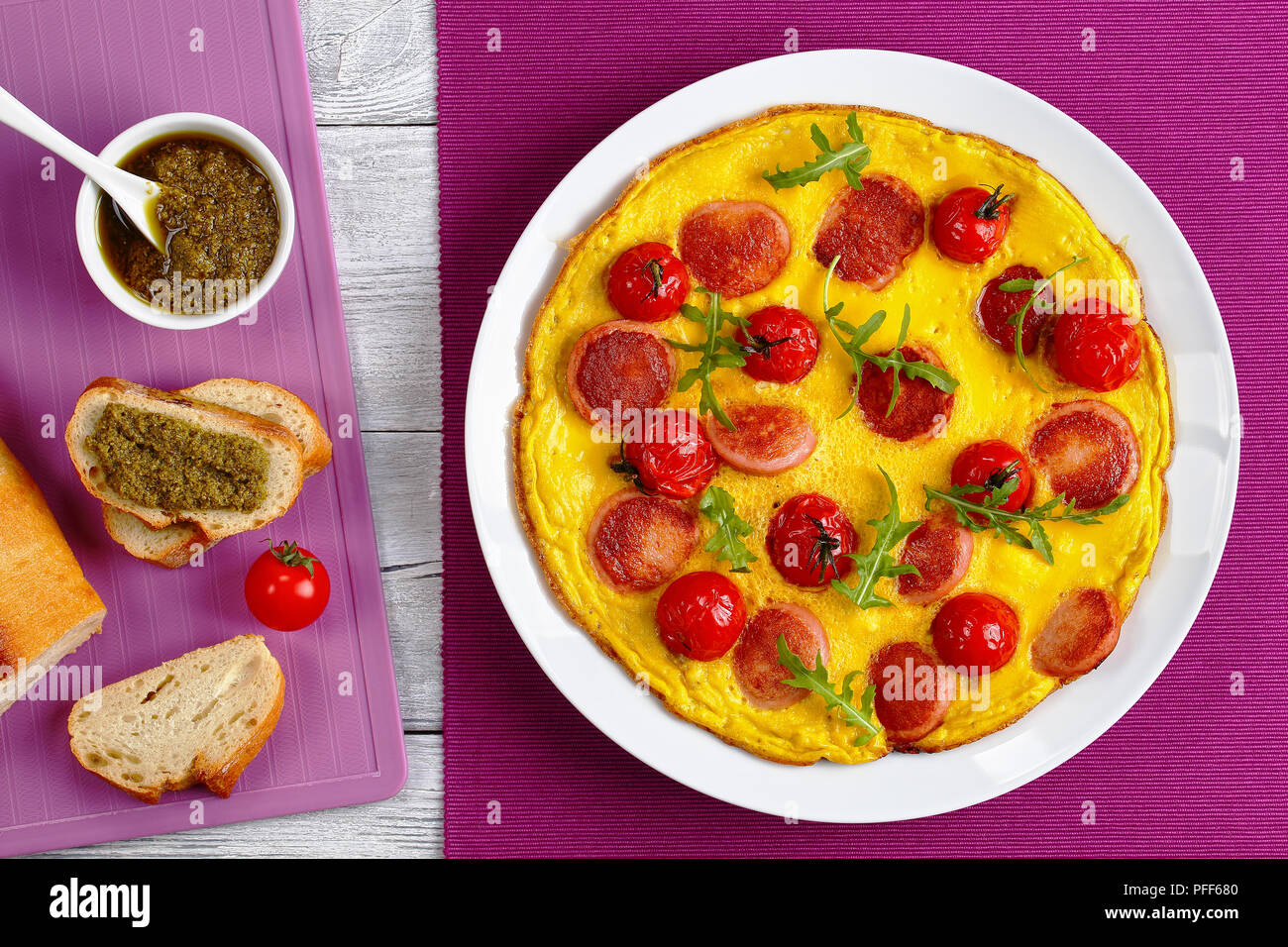 Frittata con salsiccia tagliata a fettine, interi di pomodori ciliegini e rucola fresca su bianco piatto sul tavolo in legno con pesto sandwich aperto e fren Foto Stock