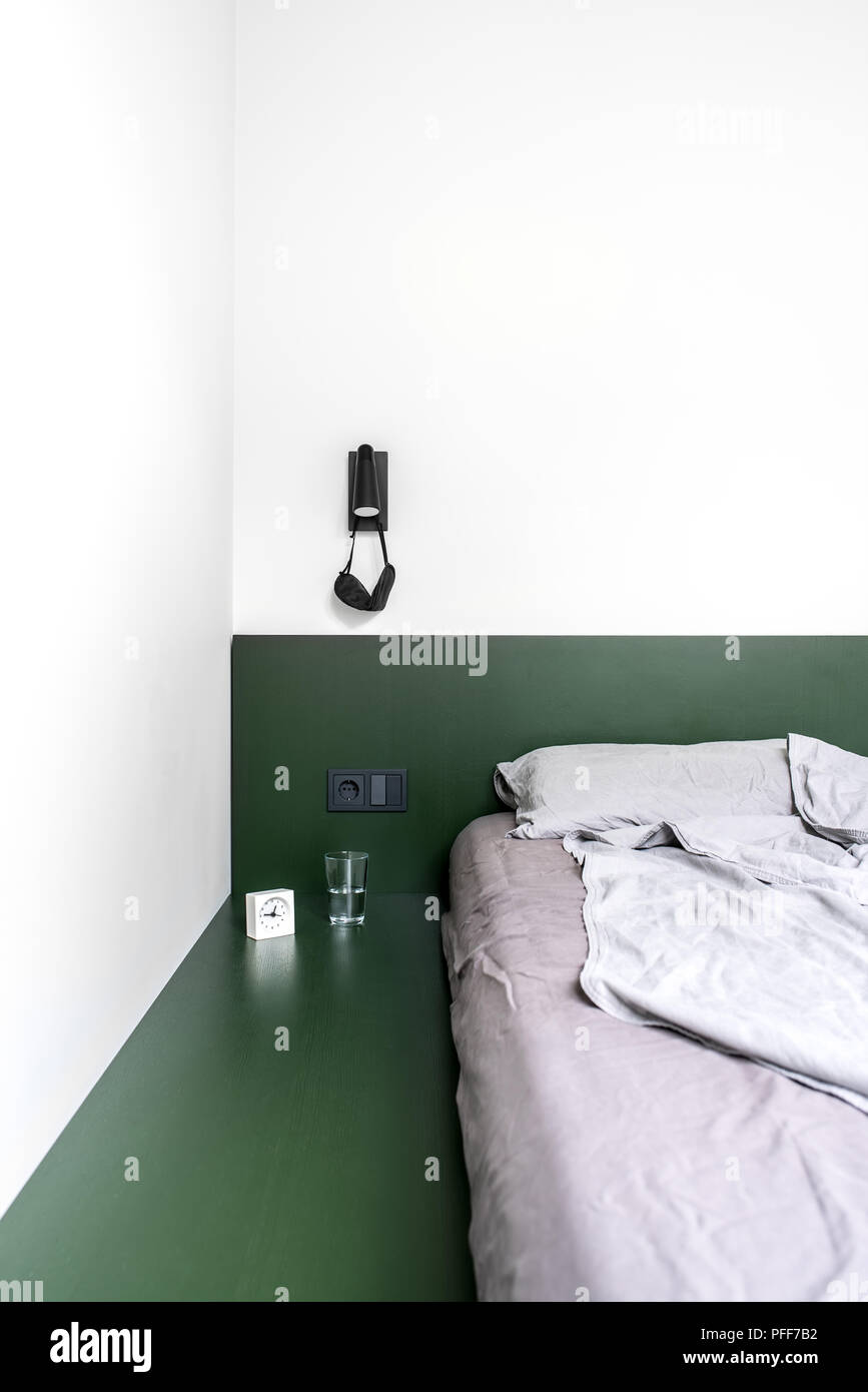 Bella camera da letto in stile moderno con pareti bianche. Vi è un letto verde pastello con lenzuola e cuscini, nero lampada a parete con appeso un occhio maschera, Foto Stock