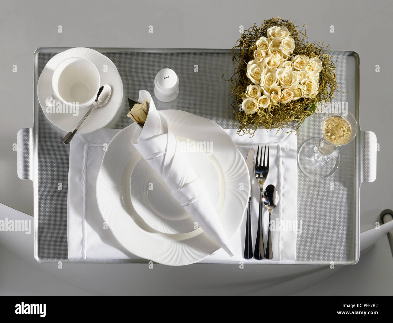 Vassoio adagiato con posate, due piastre su tela Matt, una rosa bianca, sale shaker, tazza di caffè sui coaster, bicchiere di champagne, a forma di cuore di fiori di rose testa a disposizione, vista in elevazione. Foto Stock