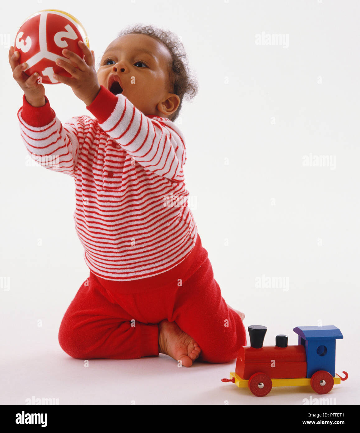 Il Toddler indossando a strisce rosse e bianche vesti, inginocchiata, sporgersi in avanti, afferrando la sfera colorata, toy train sul pavimento accanto a lei. Foto Stock