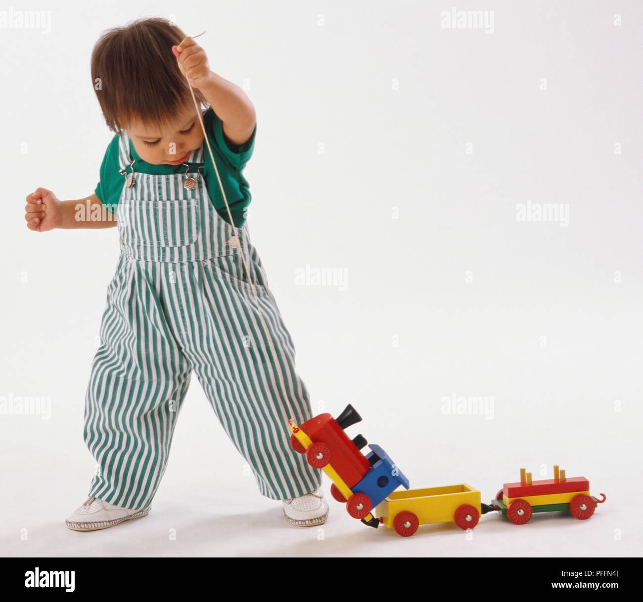 Bambino in verde e bianco striato, salopette da piedi, tirando il giocattolo di legno treno lungo il pezzo di stringa. Foto Stock