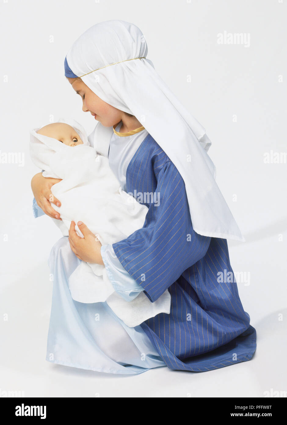 Ragazza in accappatoio blu e velo bianco inginocchiato con il giocattolo bambino avvolto in coperte bianche. Foto Stock