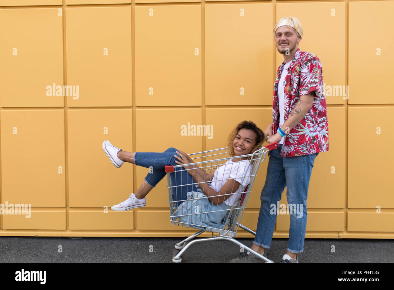 Immagine di giovani amici sorridente su parete gialla. Buon divertimento con il carrello della spesa. Foto Stock