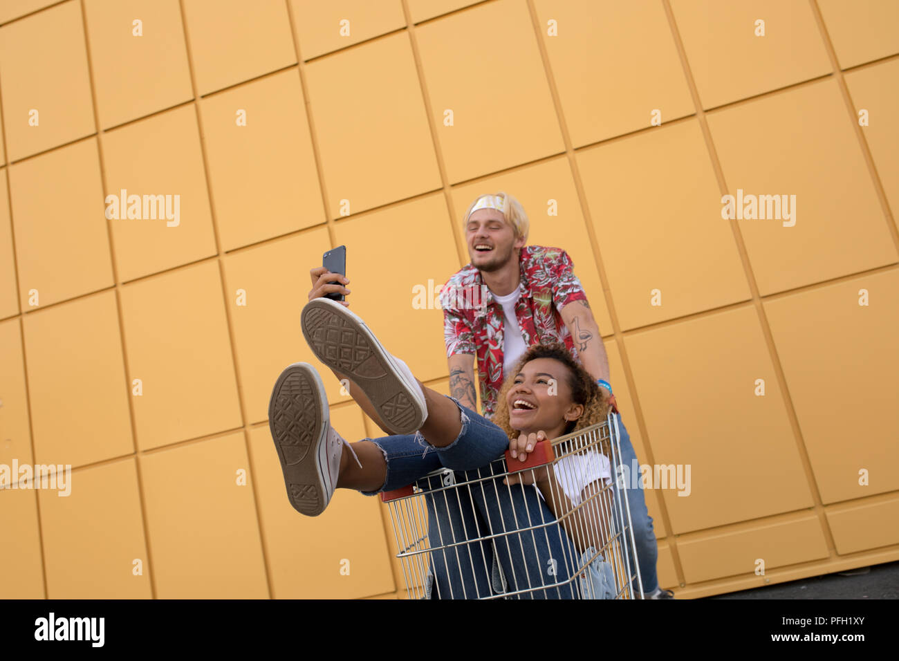 Immagine di giovani amici sorridente su parete gialla. Buon divertimento con il carrello della spesa e fare selfie. Foto Stock