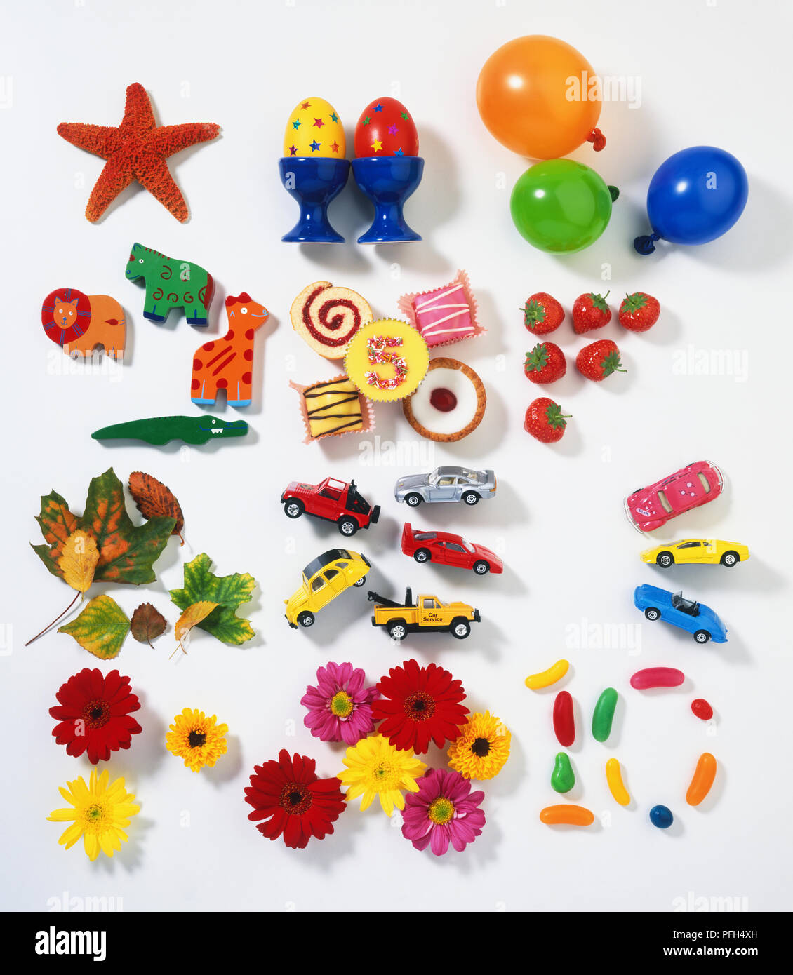 Colorata selezione di automobili in miniatura, animali, palloncini, torte, dolci, fragole, fiori, foglie dipinte di uova in tazze blu e una stella di mare Foto Stock