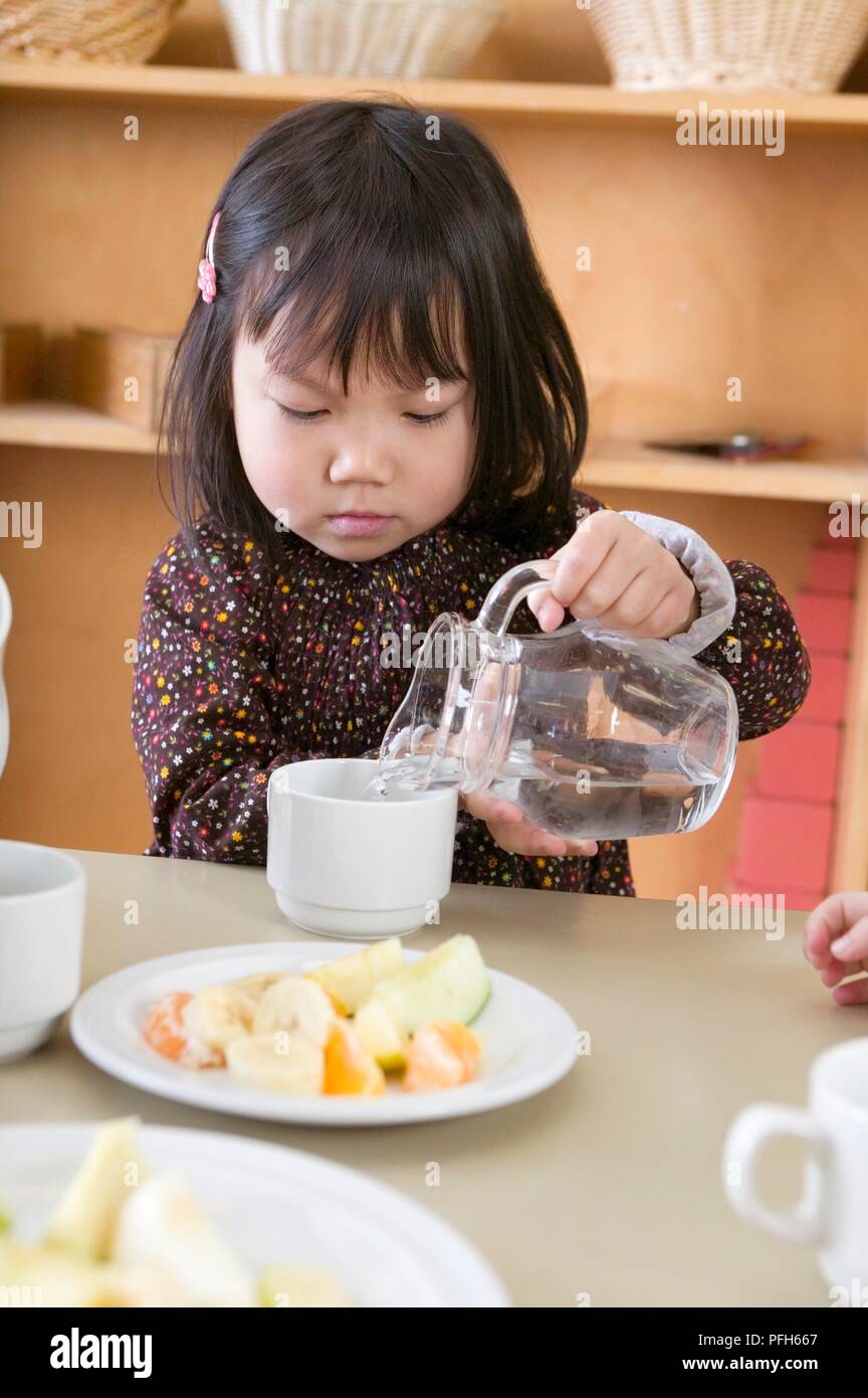Ragazza versando acqua dalla caraffa di vetro nella tazza, accanto al piatto di frutta fresca Foto Stock
