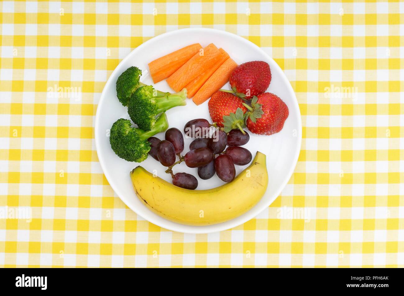 Banana, broccoli, bastoncini di carote, fragole e Uva su piastra su giallo controllato la tovaglia Foto Stock