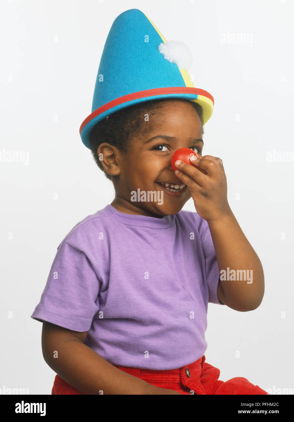 Sorridente ragazzo in blu-giallo cappello conico, rosso clown naso e t-shirt viola. Foto Stock