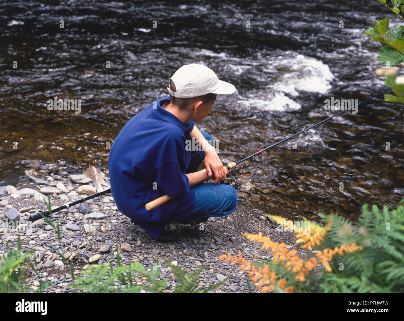 Ragazzo adolescente nel cappuccio accovacciato dal bordo del fiume casting canna da pesca in acqua, vista laterale Foto Stock