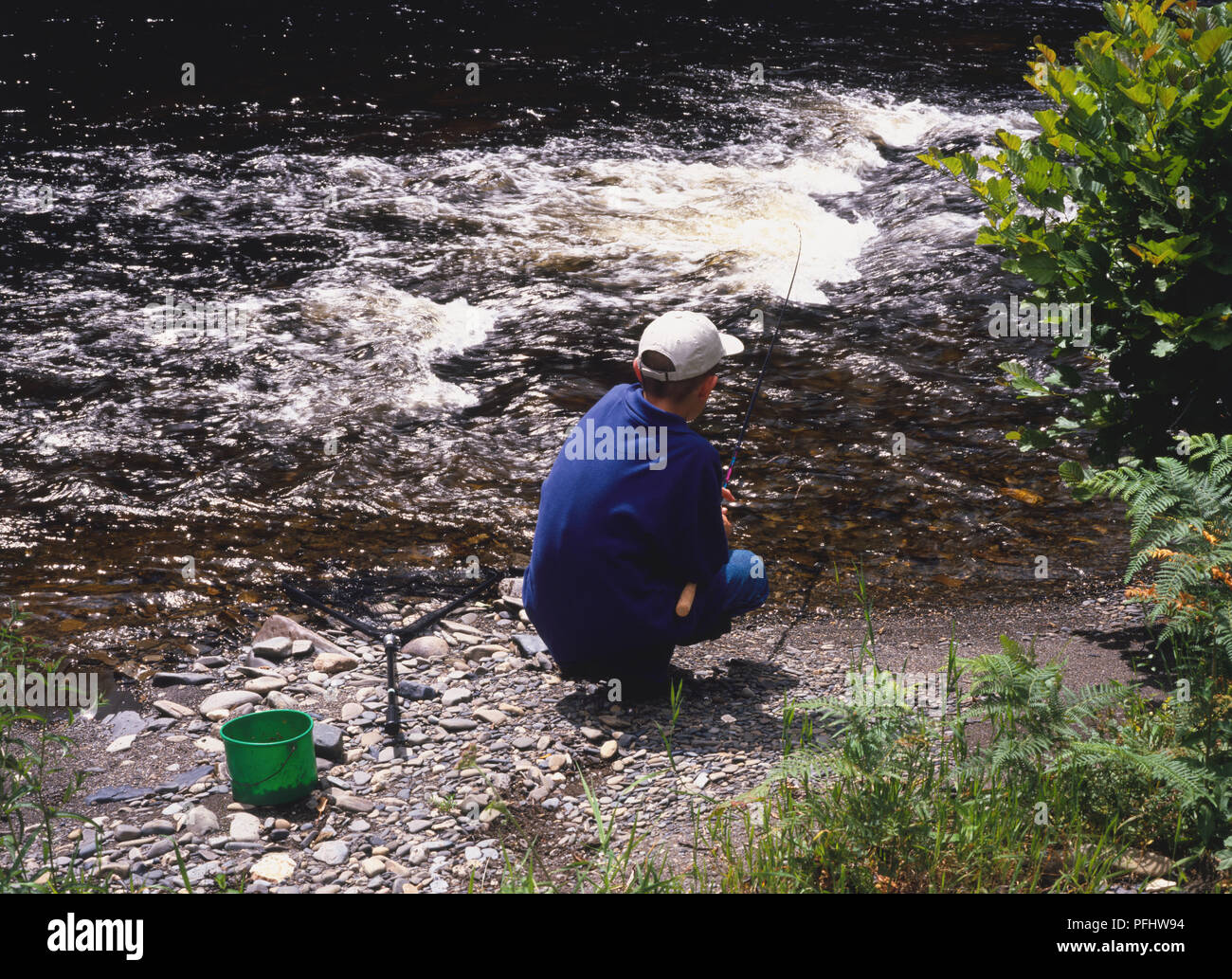 Ragazzo adolescente nel cappuccio accovacciato dal bordo del fiume casting canna da pesca in acqua, vista posteriore Foto Stock