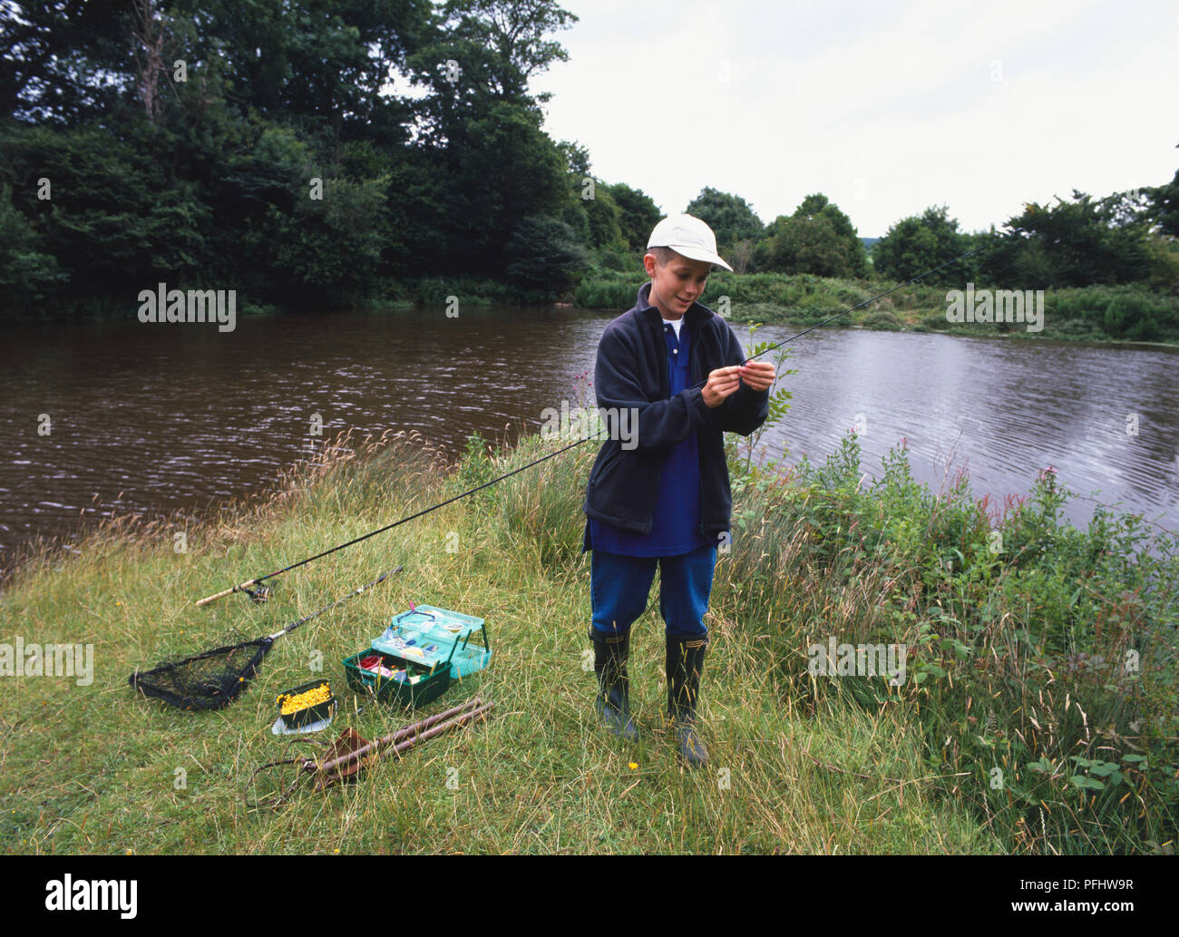 Ragazzo adolescente nel cappuccio in piedi sul prato di fronte fiume di threading linea di pesca attraverso gli anelli di asta, vista frontale Foto Stock