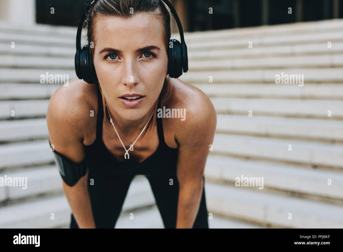 donna in forma fisica che si allena all'aperto piegandosi in avanti con le mani sulle ginocchia. donna in forma fisica che indossa le cuffie wireless per rilassarsi durante l'allenamento. Foto Stock