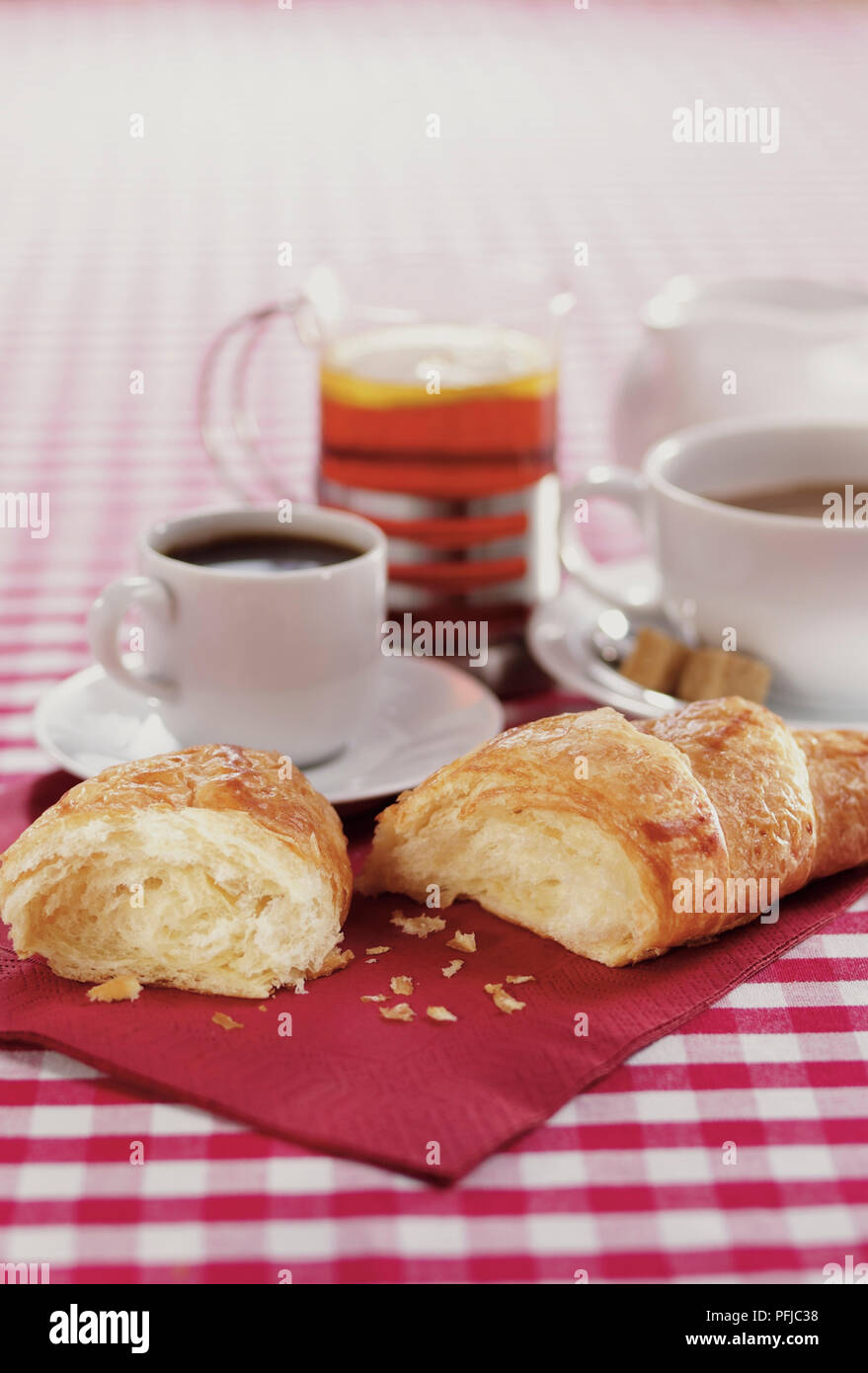 Croissant rotto in metà, una tazza di caffè espresso, una tazza di caffè e un bicchiere di tè e la brocca del latte, disteso sul tovagliolo rosso e verificata la tovaglia, close-up Foto Stock