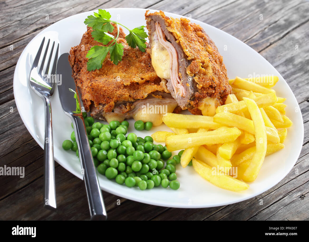 Deliziosa Cordon Bleu con patatine fritte, piselli verdi e prezzemolo sulla piastra sul vecchio legno scuro tabella d'argento con coltello e forchetta, autentica ricetta, vicino- Foto Stock