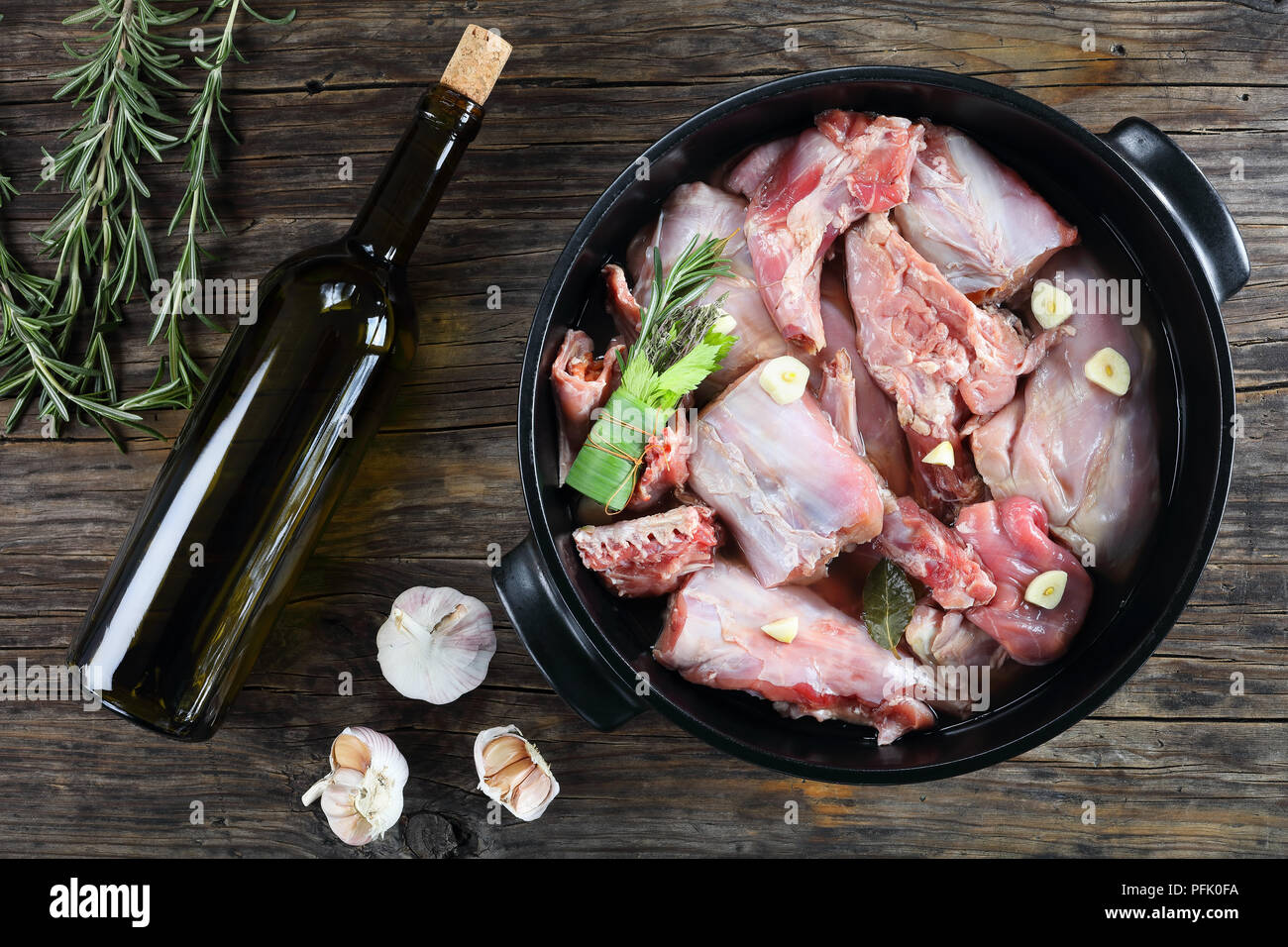 Pezzi di coniglio crudo marinato con vino bianco, aglio e il mazzetto di erbe aromatiche o bouquet garni in nero teglia sul tavolo con bottiglia di vino Foto Stock
