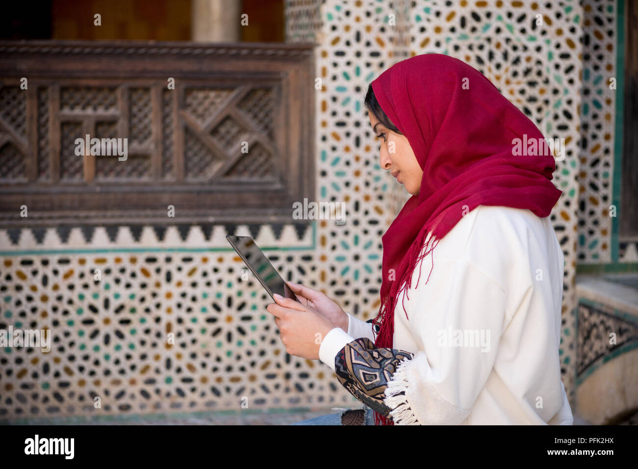 Donna musulmana lavorando su tablet in abbigliamento tradizionale con velo di colore rosso sulla sua testa accanto a un tradizionale arabesque parete marocchino Foto Stock