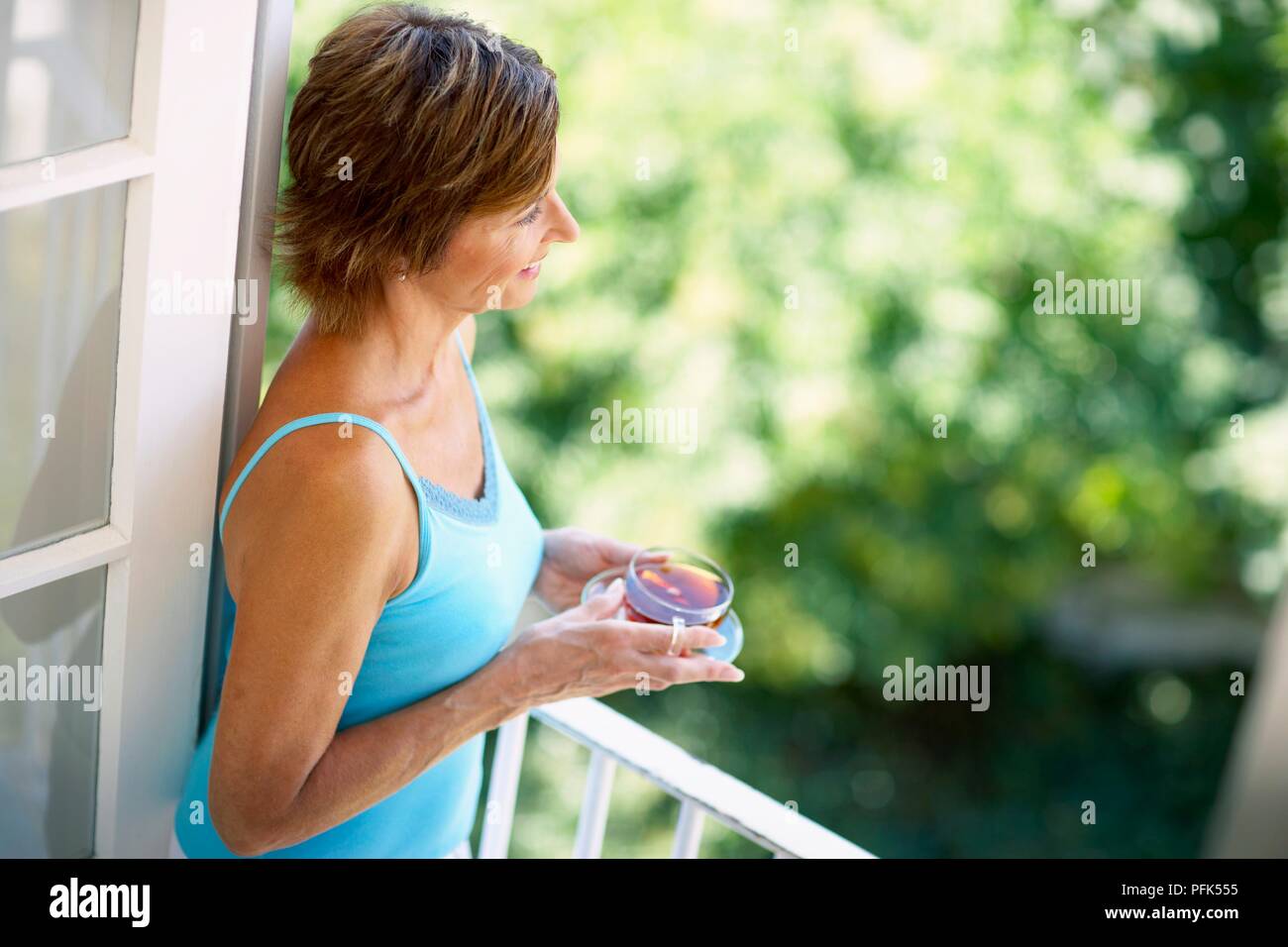 Donna giubbotto blu in piedi sul balcone, tenendo la coppa in vetro e piattino contenente tè nero Foto Stock