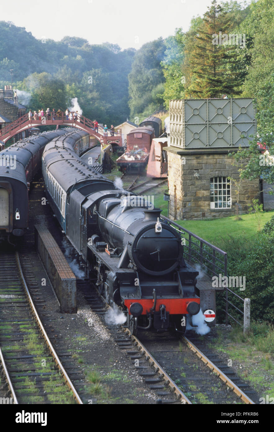 Gran Bretagna, Inghilterra, North Yorkshire, anteriore del treno a vapore che passa un altro treno durante il viaggio attraverso la campagna, la gente camminare attraverso il Footbridge in background. Foto Stock