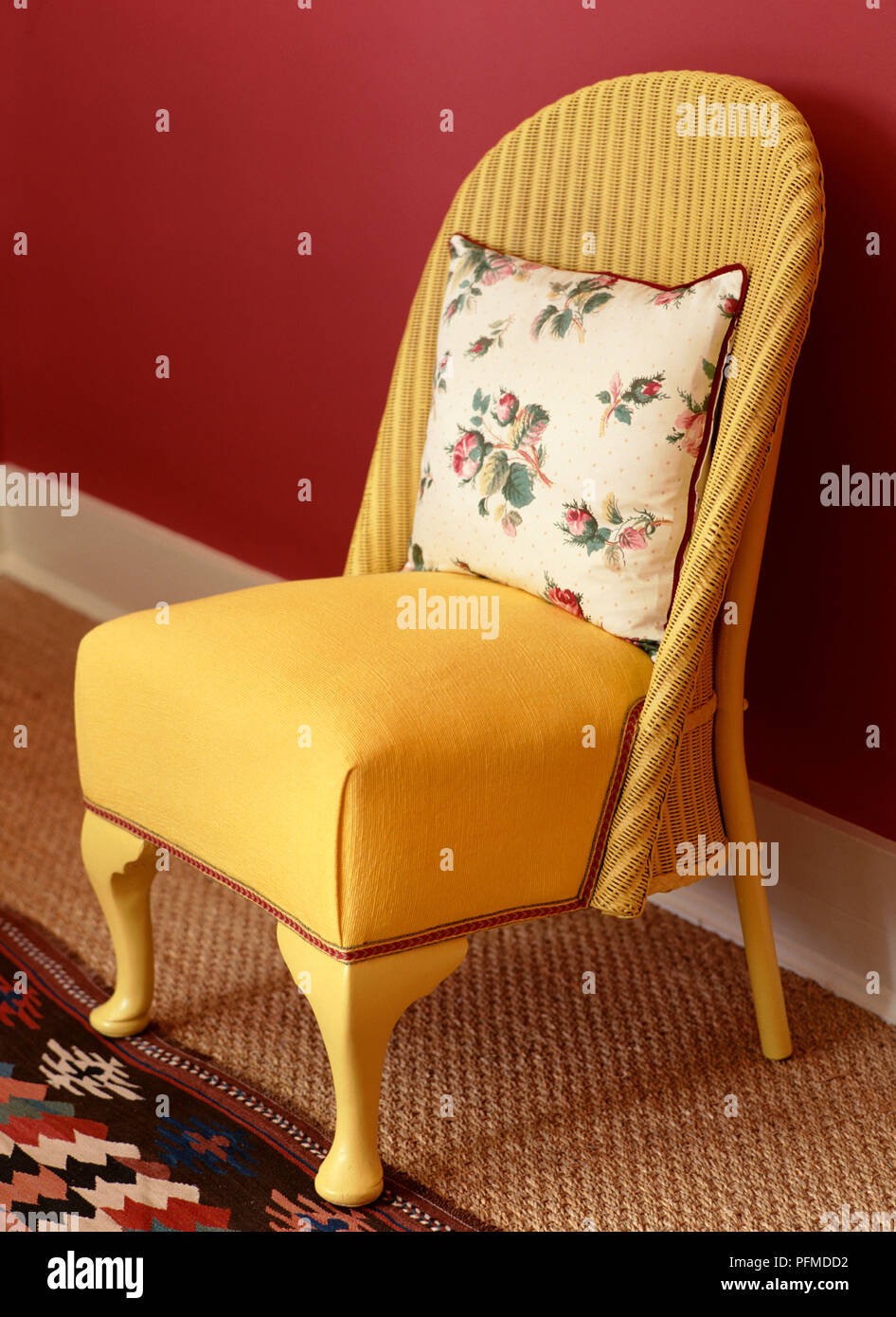Telaio giallo sedia, rosso-refilato crema cuscino fiorito in appoggio contro schienale, stuoie e modellato tappeto sul pavimento, parete rossa dietro. Foto Stock