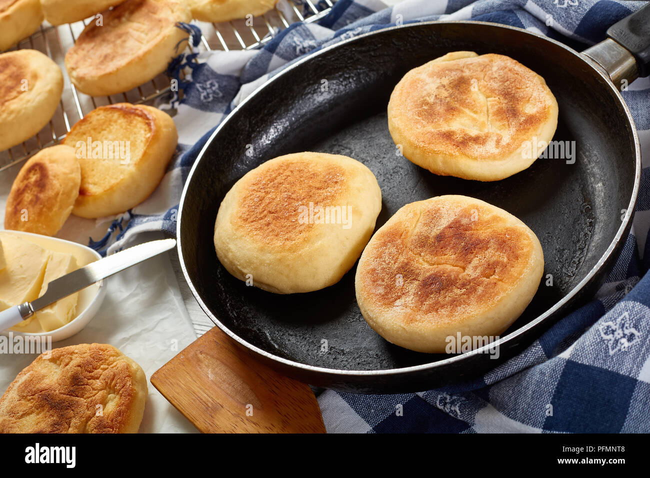Cooking per colazione - cottura muffin inglesi su una padella e sono agghiaccianti su una griglia su un tavolo di legno con carta asciugatutto, burro fresco e coltello, visualizzare fr Foto Stock