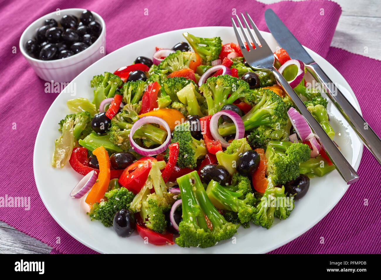 Insalata di broccoli con olive nere, peperoni rossi, anelli di cipolla e i semi di sesamo su una piastra bianca con la forchetta e il coltello sul tavolo di legno, sani a basso contenuto calorico Foto Stock