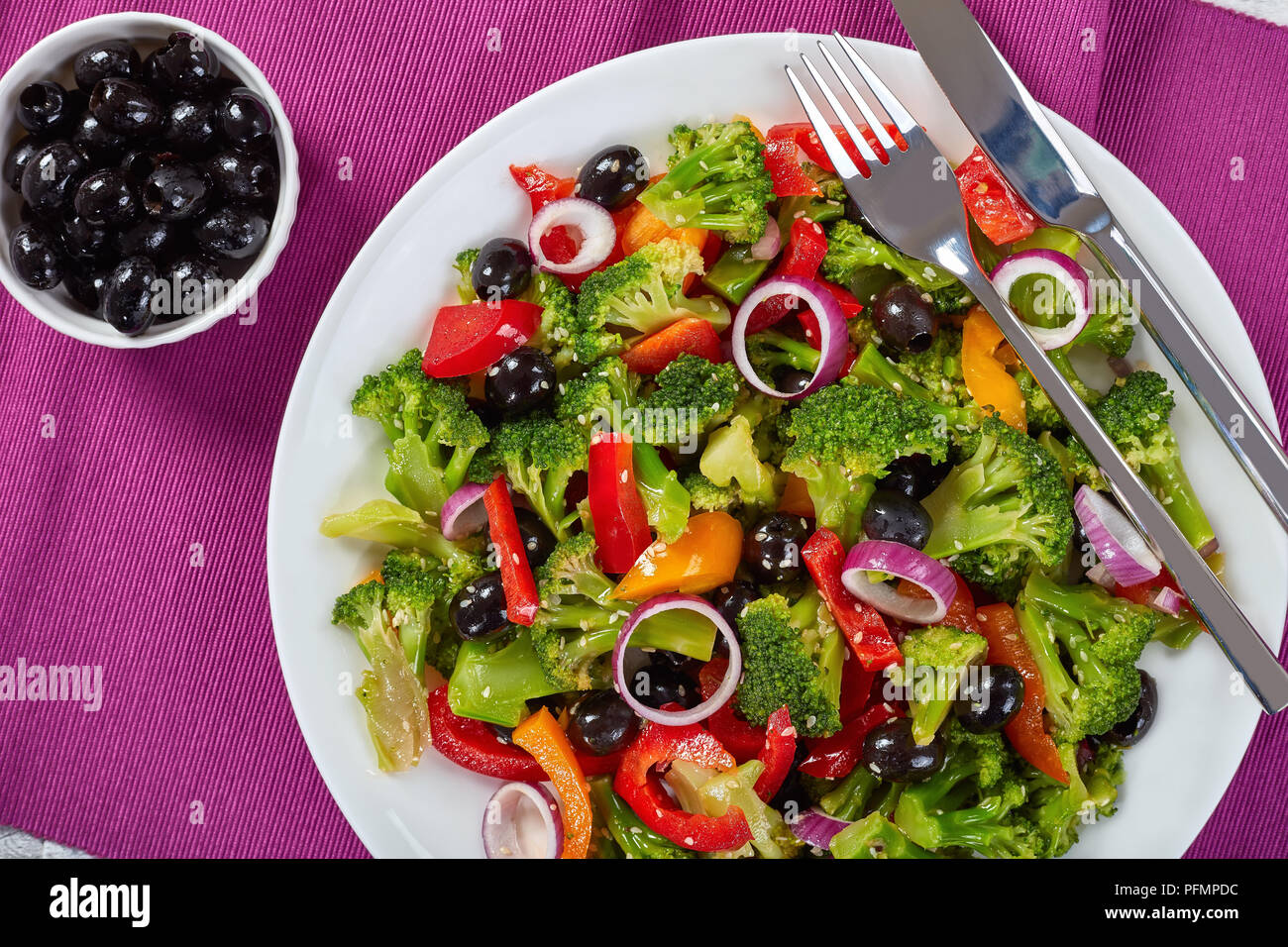Fresca Insalata di broccoli con olive nere, peperoni rossi, anelli di cipolla e i semi di sesamo su una piastra bianca con la forchetta e il coltello sul tavolo di legno, salutare l Foto Stock