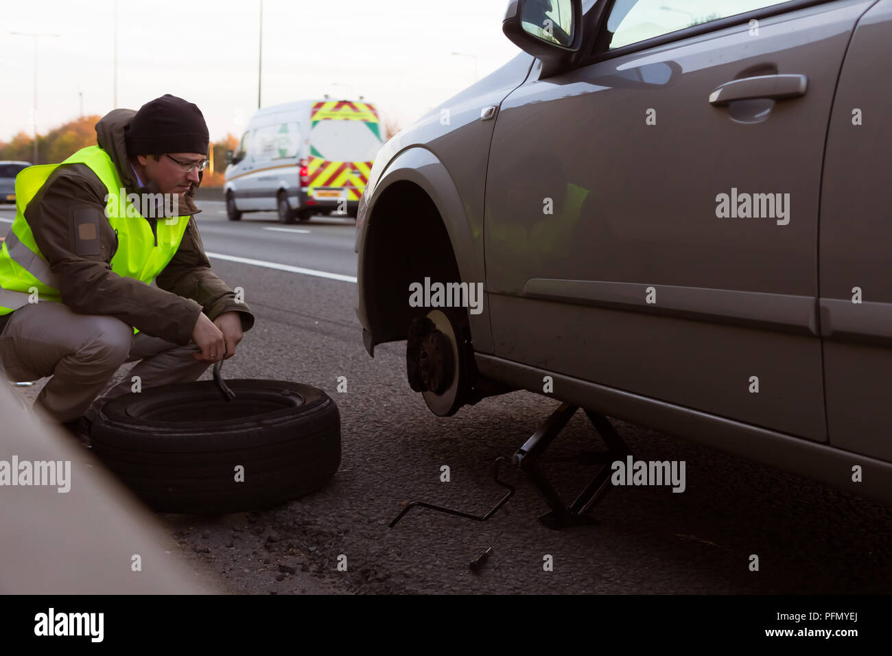 Uomo che indossa alta visibilità giacca cambia una ruota con pneumatico sgonfio per una vettura ferma in corsia di autostrada Foto Stock